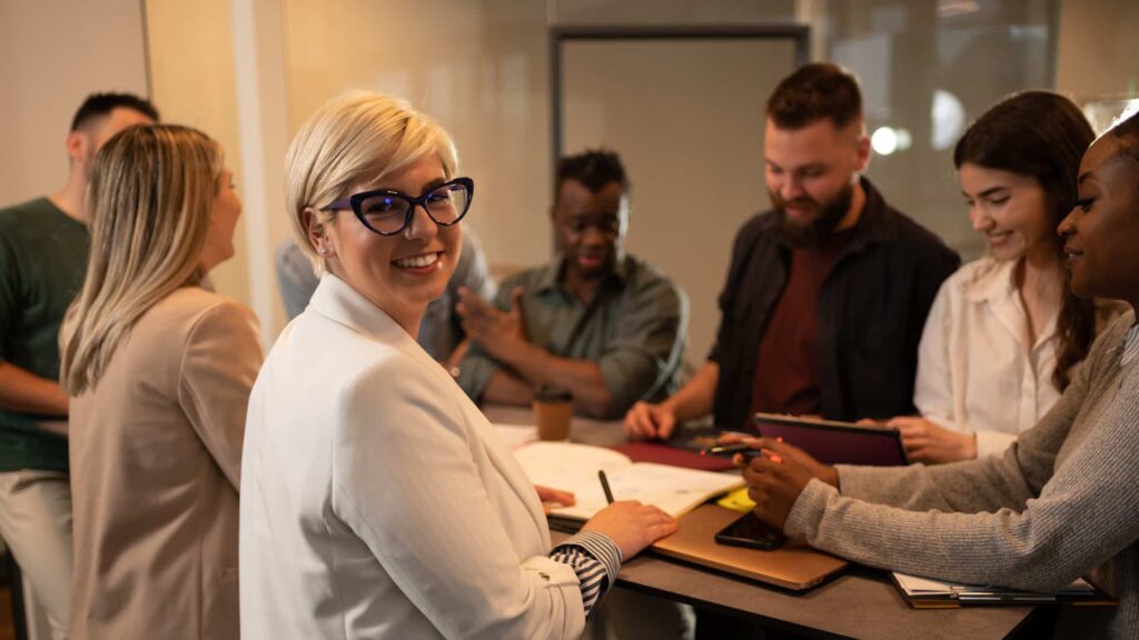 Donna sorridente a un tavolo di lavoro con altri colleghi.